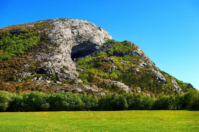 A large rocky mountain with a prominent cave opening, covered in green vegetation, rises above a lush green meadow and a line of trees under a clear blue sky.