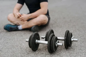 A person sitting on the ground with legs crossed, wearing a black shirt and shorts, with two dumbbells placed in front of them on a paved surface.