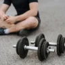A person sitting on the ground with legs crossed, wearing a black shirt and shorts, with two dumbbells placed in front of them on a paved surface.