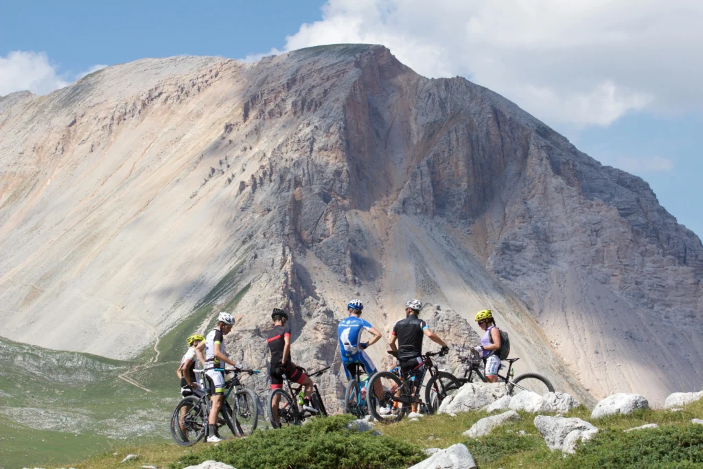 A group of five cyclists wearing helmets and colorful biking gear stand with their mountain bikes on a grassy hill, facing a large, rugged mountain with rocky slopes under a partly cloudy sky.