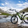 A green Pinarello electric mountain bike is parked on a rocky terrain near a flowing stream, with snow-covered mountains and green hills in the background under a bright blue sky with scattered clouds.