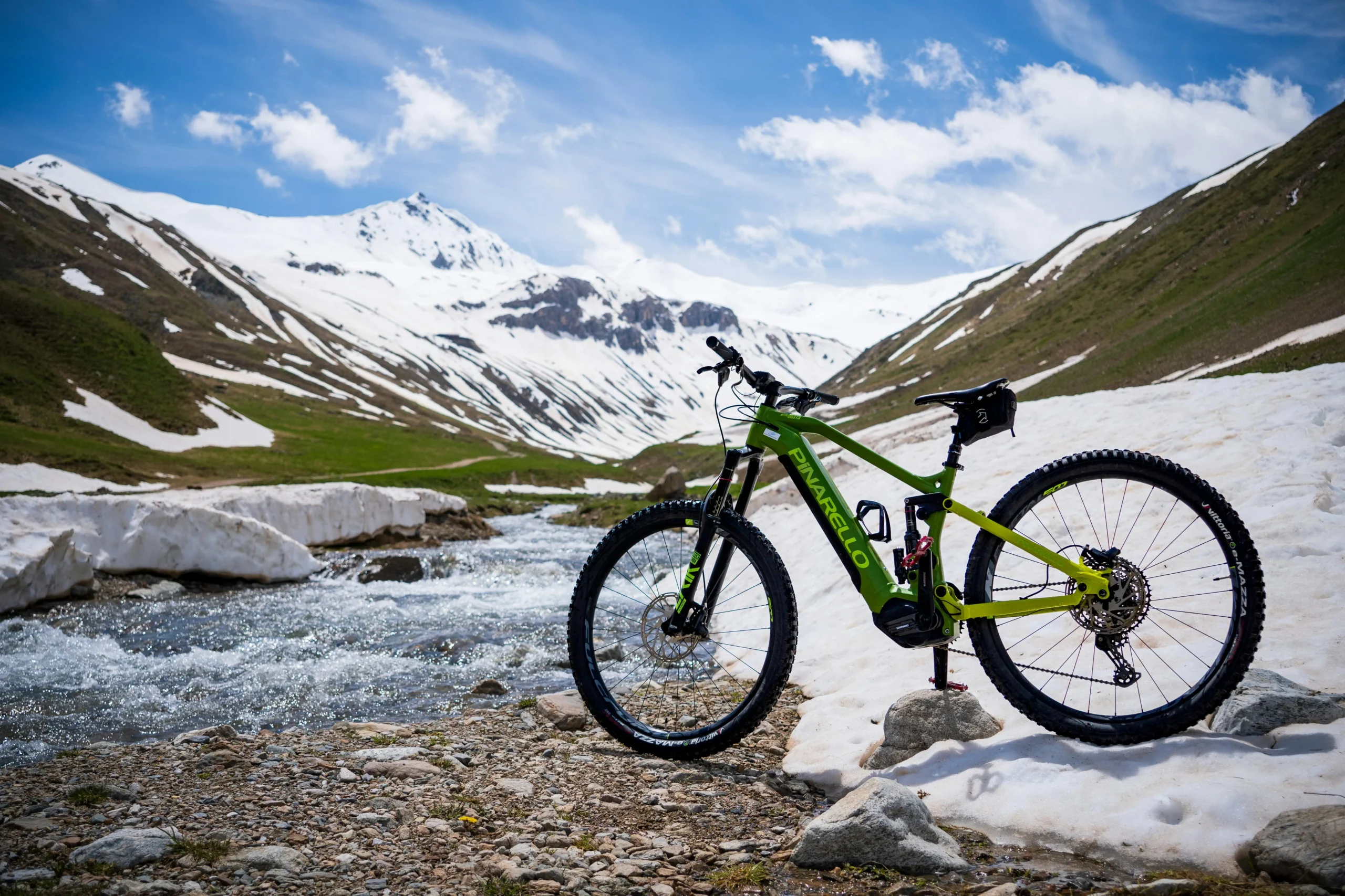A green Pinarello electric mountain bike is parked on a rocky terrain near a flowing stream, with snow-covered mountains and green hills in the background under a bright blue sky with scattered clouds.