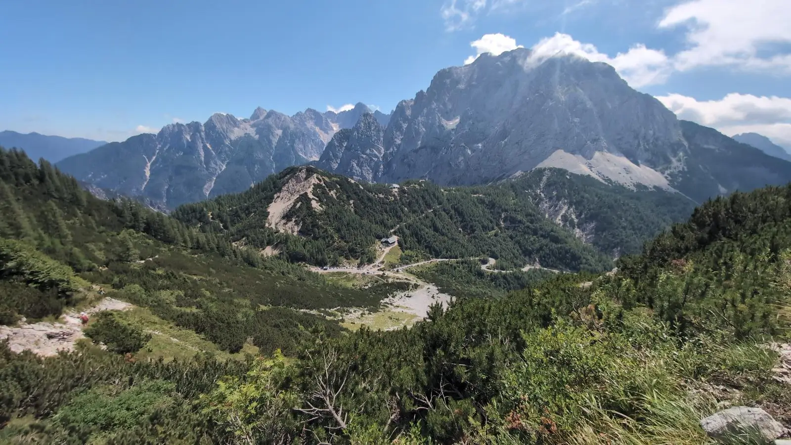 A breathtaking view of the Julian Alps in Slovenia on a clear summer day, with lush green valleys, dense coniferous forests, and dramatic limestone peaks rising sharply in the background under a deep blue sky. A small mountain hut and winding road are visible in the mid-ground.