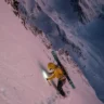 A skier in a yellow jacket climbs a steep, snow-covered mountain slope at dawn or dusk, using a headlamp to illuminate the path ahead. The skier carries skis on their back and holds an ice axe in one hand. The vast, rugged mountain landscape stretches into the distance under a gradient sky transitioning from pink to blue.