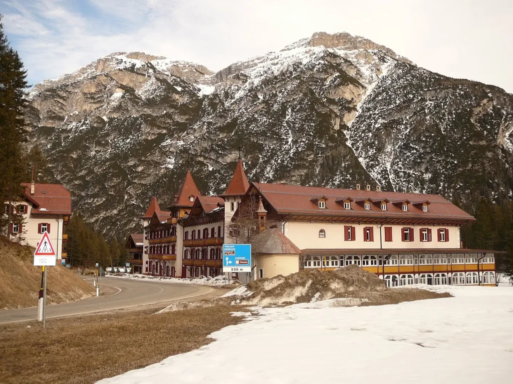 A grand, multi-story alpine hotel with steep red roofs and wooden balconies stands beside a winding road, flanked by snow-dusted ground and evergreen trees. In the background, towering snow-capped mountains rise sharply under a partly cloudy sky. Road signs in the foreground indicate directions and a pedestrian crossing.