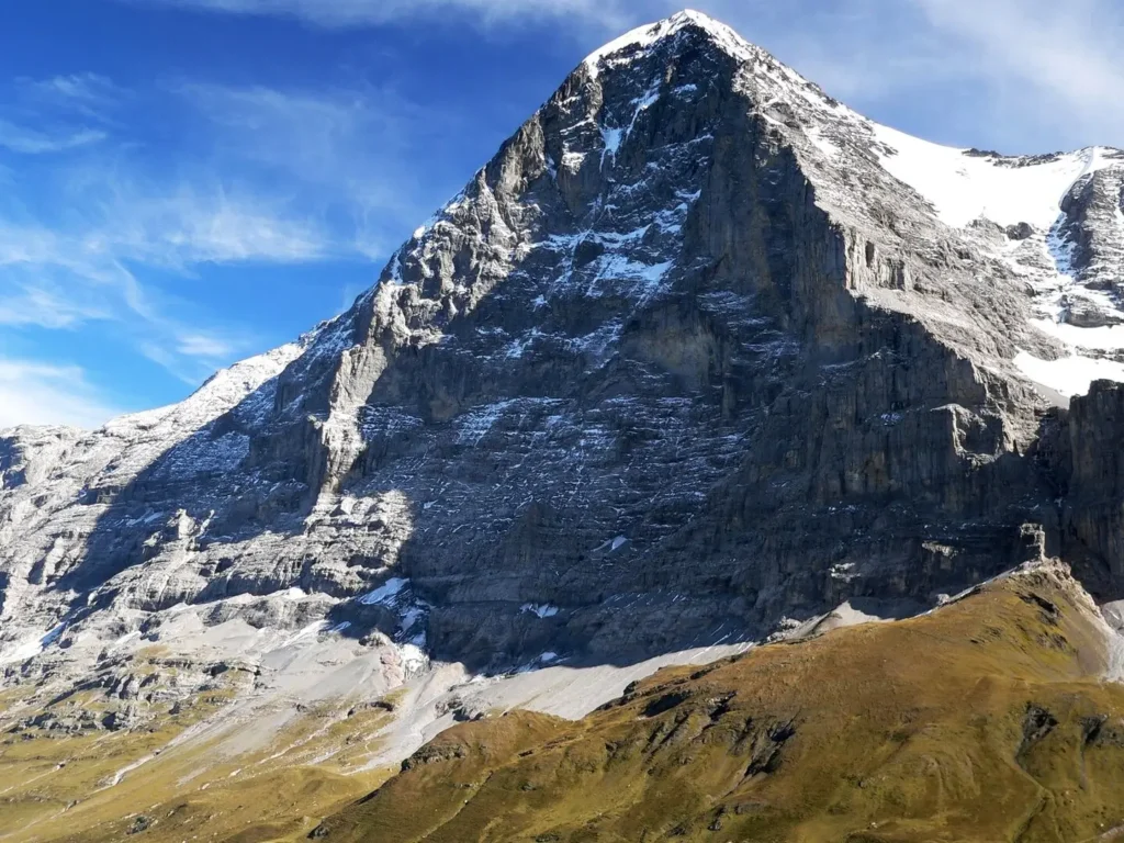 A striking color photograph of the massive, near-vertical north face of the Eiger (Eiger Nordwand) in the Swiss Alps. The imposing rock wall rises thousands of feet, streaked with snow and ice, under a clear blue sky. The steep, foreboding face dominates the frame, illustrating why it is known as one of the most challenging and dangerous climbs in the world.