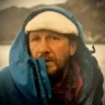 A bearded man wearing a light-colored knit cap and a blue hooded jacket pulled up around his face, standing outdoors near a body of water with a blurred natural landscape in the background.