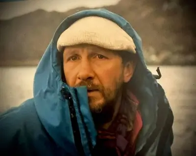 A bearded man wearing a light-colored knit cap and a blue hooded jacket pulled up around his face, standing outdoors near a body of water with a blurred natural landscape in the background.