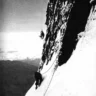 A dramatic black-and-white photograph of two climbers on the near-vertical northeast face of the Eiger. One climber is high above, dangling precariously from a rope against the sheer rock wall, while the second climber, far below, stands on a steep snow slope holding the rope, looking up at their partner. The vast drop and clouds below emphasize the extreme exposure of the Eiger Nordwand.