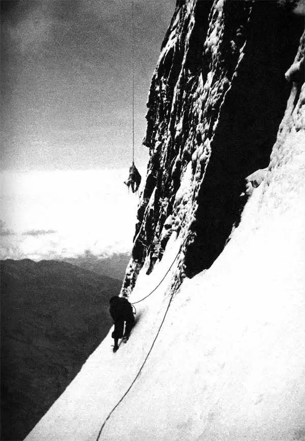 A dramatic black-and-white photograph of two climbers on the near-vertical northeast face of the Eiger. One climber is high above, dangling precariously from a rope against the sheer rock wall, while the second climber, far below, stands on a steep snow slope holding the rope, looking up at their partner. The vast drop and clouds below emphasize the extreme exposure of the Eiger Nordwand.
