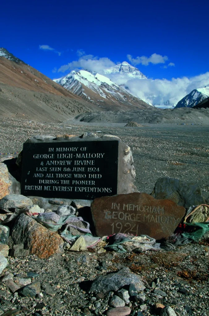 A black memorial plaque on a rocky cairn at Everest North Base Camp, reading “IN MEMORY OF GEORGE LEIGH-MALLORY & ANDREW IRVINE LAST SEEN 8th JUNE 1924 AND ALL THOSE WHO DIED DURING THE PIONEER BRITISH MT. EVEREST EXPEDITIONS.” A smaller stone in front bears the carved inscription “IN MEMORIAM GEORGE MALLORY 1924.” Mount Everest’s snow-capped north face rises prominently under a clear blue sky in the background, surrounded by barren high-altitude terrain and prayer flags.