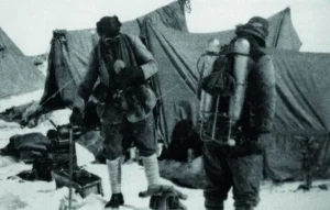 Black-and-white photograph from the 1924 British Mount Everest Expedition showing George Mallory (left) and Andrew “Sandy” Irvine (right) at or near the North Col. Both are dressed in heavy layered wool clothing, hobnail boots, and early climbing gear, carrying ice axes and wearing wide-brimmed hats. They are roped together on snowy, rocky terrain with tent fabric visible behind them. This is the last known photograph of the pair before their fatal summit attempt.