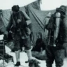 Black-and-white photograph from the 1924 British Mount Everest Expedition showing George Mallory (left) and Andrew “Sandy” Irvine (right) at or near the North Col. Both are dressed in heavy layered wool clothing, hobnail boots, and early climbing gear, carrying ice axes and wearing wide-brimmed hats. They are roped together on snowy, rocky terrain with tent fabric visible behind them. This is the last known photograph of the pair before their fatal summit attempt.