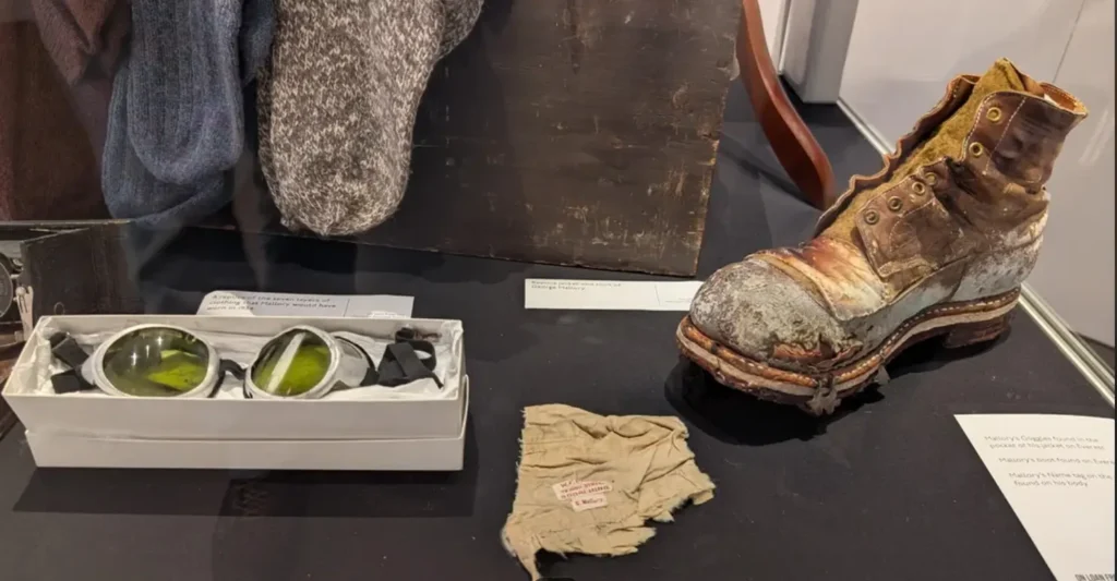 Museum display case featuring artifacts from Mount Everest, including a heavily weathered and deteriorated leather climbing boot with thick sole and metal eyelets, a pair of green-tinted glacier goggles in a white box, a tattered fabric name tag labeled "A. Mallory" and "G. Mallory," and hanging wool socks. Placards describe the items as belonging to Andrew "Sandy" Irvine and George Mallory from their 1924 Everest expedition, with the boot and goggles found on Mallory's body and the name tag recovered nearby.