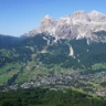 Aerial view of a picturesque alpine village nestled in a lush green valley, surrounded by dense forests and rolling meadows, with majestic snow-capped mountains rising dramatically in the background under a clear blue sky.