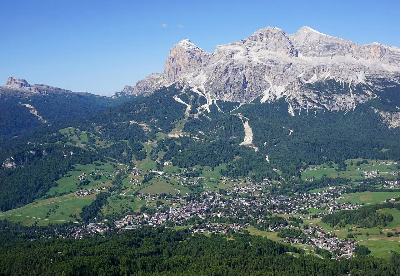 Aerial view of a picturesque alpine village nestled in a lush green valley, surrounded by dense forests and rolling meadows, with majestic snow-capped mountains rising dramatically in the background under a clear blue sky.