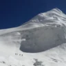 A group of climbers ascends a steep, snow-covered slope toward a towering, jagged mountain peak under a clear blue sky. The mountain features prominent ice cliffs and crevasses, with the climbers appearing small against the vast, icy landscape.
