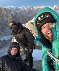 Three climbers in heavy winter gear pose for a selfie on a rocky, snow-covered ridge with a dramatic backdrop of jagged, snow-capped mountain peaks under a clear blue sky.