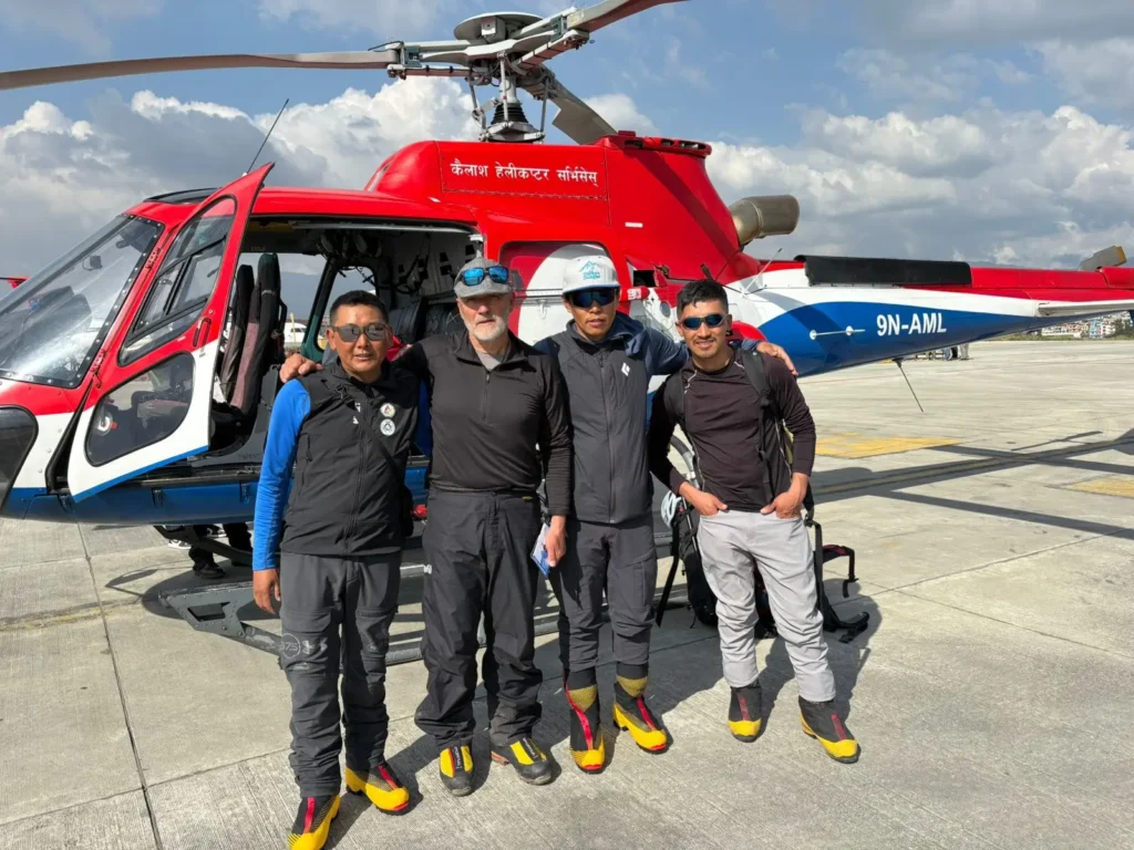 Four mountaineers in technical gear, including high-altitude boots and jackets, stand smiling in front of a red and blue helicopter with the registration "9N-AML." The helicopter door is open, and the group appears ready for a high-altitude expedition under a partly cloudy sky.
