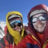 Two smiling climbers in heavy mountaineering gear (one in a bright yellow hooded jacket, the other in red and purple) take a selfie high on a snowy mountain ridge under a deep blue sky, both wearing helmets, glacier glasses, and frost-covered faces, with distant peaks visible behind them.