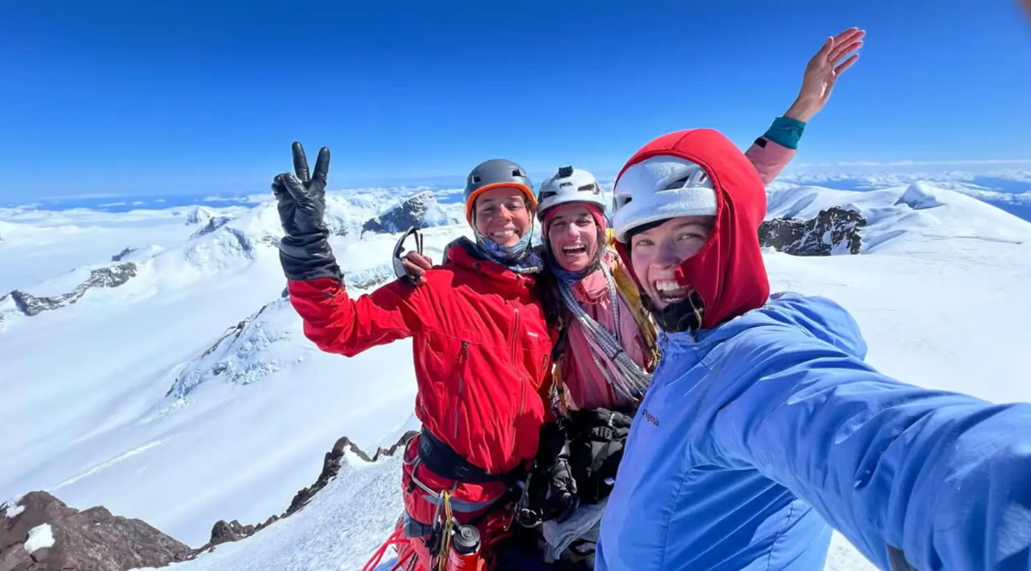 Three smiling climbers in helmets and mountaineering gear pose for a selfie on a snowy mountain summit. One makes a peace sign, another waves, and the third takes the photo. The background features a stunning expanse of snow-covered peaks under a clear blue sky.