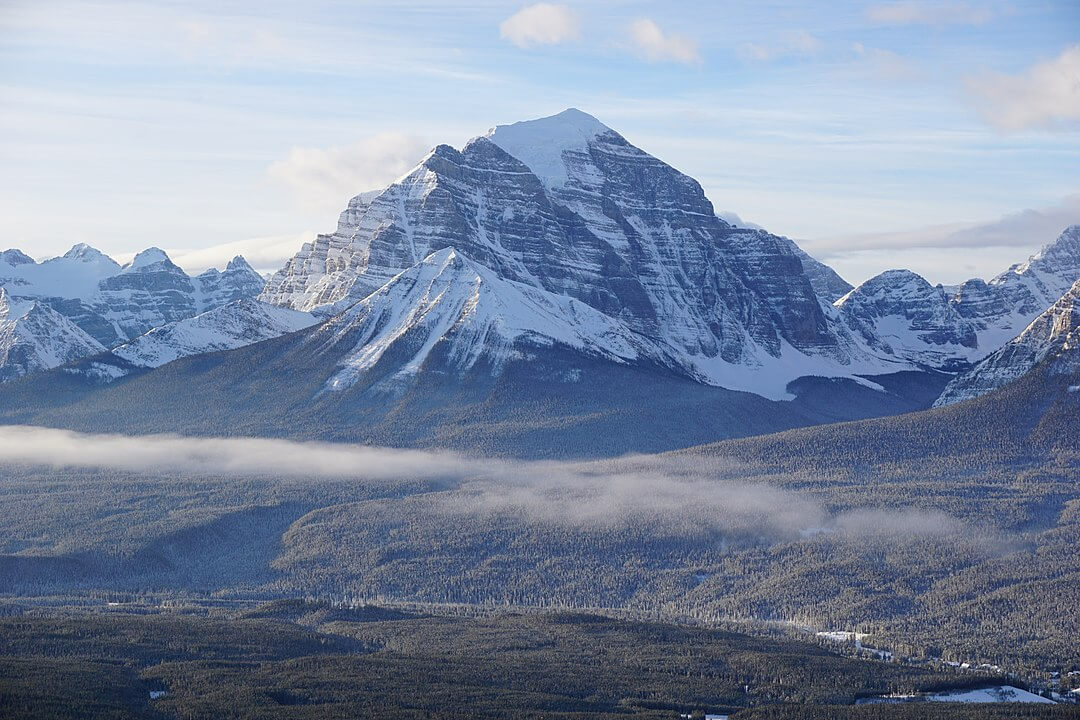Mount Temple rising above Banff National Park in winter, its snow-covered limestone face towering over forested valleys under a clear blue sky.