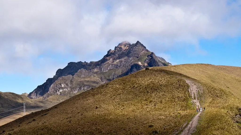 A dramatic high-altitude landscape featuring a steep, grassy hillside with a narrow dirt trail winding down toward the viewer. In the distance, a rugged, dark, jagged mountain peak rises sharply into a partly cloudy sky, its summit partially shrouded in low clouds. A small group of hikers is visible far down the trail near the bottom right, emphasizing the vast scale of the scene.