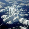 Aerial view of rugged, snow-capped mountain peaks bathed in sunlight, with sharp ridges and deep valleys casting dramatic shadows. Dark evergreen forests blanket the lower slopes, contrasting against the bright white snow cover under a clear blue sky.