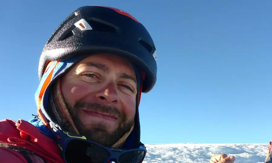 A close-up selfie of a bearded mountaineer smiling squint-eyed in bright sunlight, wearing a black climbing helmet, colorful buff pulled up over his mouth and nose, a red down jacket, and sunglasses hanging from his neck. The background shows a vast, flat expanse of snow under a clear deep blue sky, suggesting a high-altitude summit or plateau.