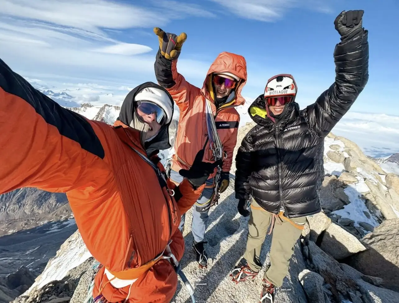 Three mountaineers celebrate on a snowy alpine summit, taking a selfie against a stunning backdrop of jagged peaks and blue sky. The person on the left, in an orange jacket, holds the camera while making a shaka hand gesture. The middle climber, also in orange, flashes a big smile, and the climber on the right, wearing a black puffy jacket and white helmet, raises a triumphant fist. All are equipped with climbing gear, helmets, and sunglasses, standing on rocky, snow-dusted ground high above the clouds.
