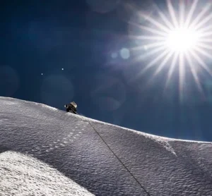 A lone mountaineer in full high-altitude gear climbs a steep, snow-covered ridge under a deep blue sky. The bright sun dominates the upper right, creating intense lens flares and starburst effects, while a fixed rope trails down the slope behind the climber, emphasizing the extreme exposure and altitude.