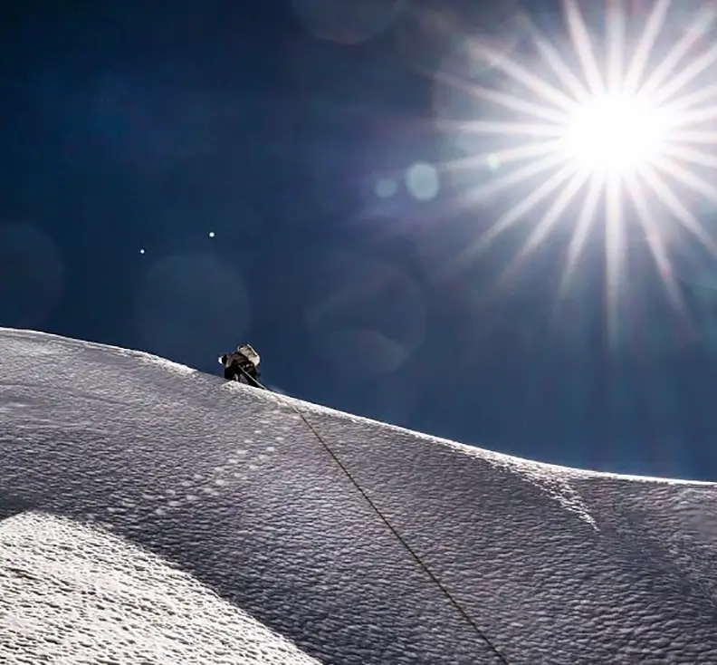 A lone mountaineer in full high-altitude gear climbs a steep, snow-covered ridge under a deep blue sky. The bright sun dominates the upper right, creating intense lens flares and starburst effects, while a fixed rope trails down the slope behind the climber, emphasizing the extreme exposure and altitude.