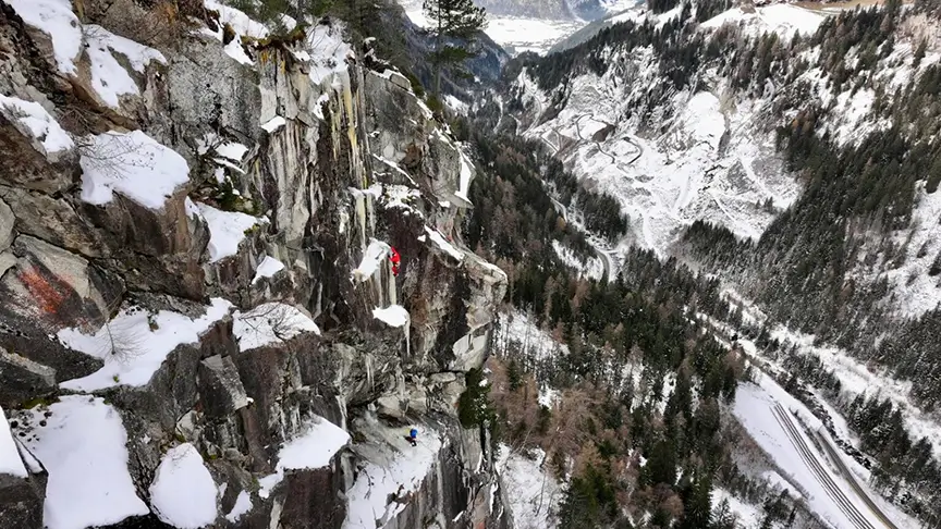 A daring climber in a red jacket scales a steep, snow-dusted granite rock face high above a deep alpine valley. Far below, a winding mountain road snakes through snow-covered evergreen forests, with distant peaks and a frozen valley floor visible under a cold, overcast sky. The sheer drop and vast scale emphasize the extreme exposure of the climb.