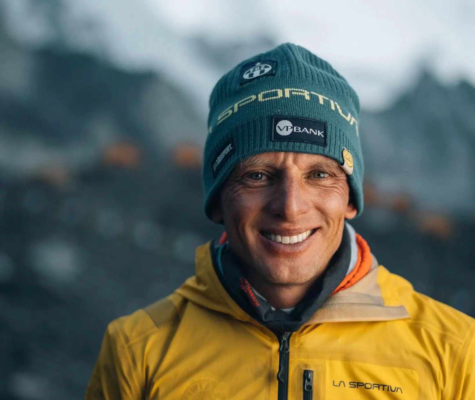 A smiling mountaineer poses for a close-up portrait in a snowy alpine environment, looking directly at the camera with a relaxed expression. He wears a teal La Sportiva beanie with patches including "VP Bank" and "La Sportiva," an orange neck gaiter, and a bright yellow La Sportiva jacket. The blurred background shows a misty, snow-covered mountainous landscape.