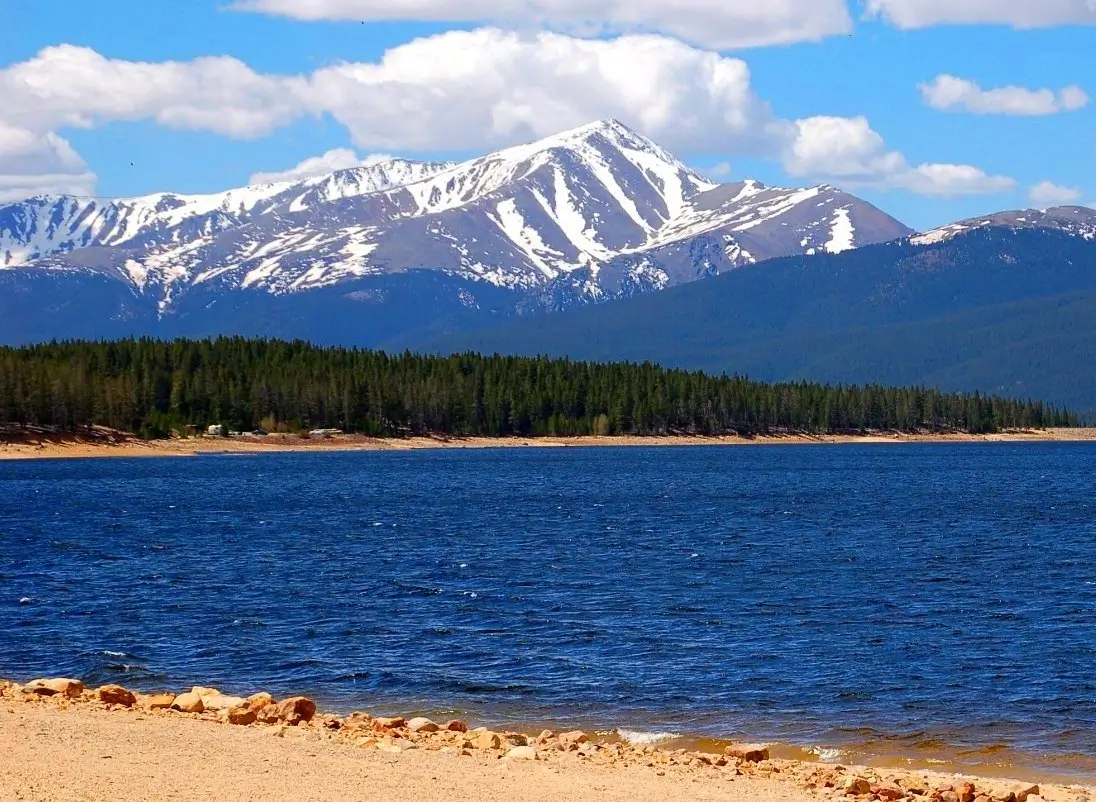 A serene alpine lake with deep blue water gently lapping at a rocky, sandy shoreline in the foreground. A dense forest of dark green evergreen trees lines the opposite shore, leading up to a majestic snow-capped mountain peak rising prominently against a bright blue sky dotted with scattered white clouds. The scene captures a peaceful, picturesque natural landscape on a clear day.