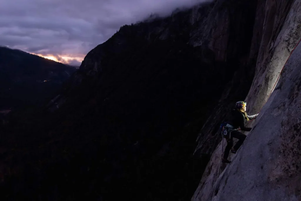 A climber wearing a headlamp ascends a sheer granite wall at night on El Capitan in Yosemite Valley. The headlamp casts a small pool of light on the rock while the vast darkness of the valley drops away thousands of feet below, with faint distant lights visible far beneath a twilight sky.