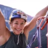 A smiling female climber in a helmet and harness raises both arms in triumphant celebration at the summit of a big wall route, likely El Capitan, at sunrise or sunset. She is brightly lit against the deep shadow of the rock face behind her, with climbing gear and ropes hanging from her harness.