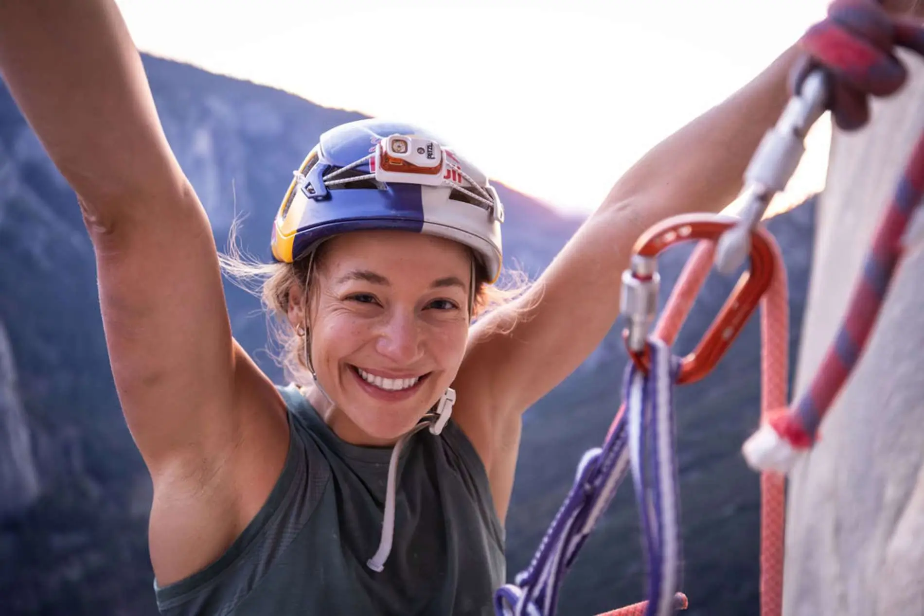 A smiling female climber in a helmet and harness raises both arms in triumphant celebration at the summit of a big wall route, likely El Capitan, at sunrise or sunset. She is brightly lit against the deep shadow of the rock face behind her, with climbing gear and ropes hanging from her harness.